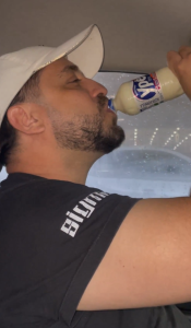 Homem usando boné branco e camiseta preta com a inscrição 'BRUNO' bebendo de uma garrafa de detergente Ypê de Côco, em um ângulo de baixo para cima