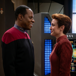 O Comandante Benjamin Sisko, à esquerda em uniforme Starfleet, e a Major Kira Nerys, à direita em uniforme vinho, conversam e sorriem na estação orbital Deep Space Nine, com equipamento técnico e painéis iluminados em azul visíveis ao fundo.