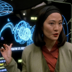 Keiko O'Brien, professora e cientista da série Star Trek DS9, em pé diante de painéis luminosos com gráficos azuis em uma sala técnica da estação. Ela usa um blazer cinza sobre uma blusa vermelha, tem cabelo preto comprido e está gesticulando com a mão direita levantada em um gesto de explicação ou demonstração. Ao fundo, telas e interfaces técnicas iluminadas compõem o cenário futurista típico da série.