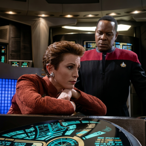 Comandante Sisko e Major Kira na cabine de comando da estação espacial Deep Space Nine. Kira, à esquerda, veste uniforme vermelha da Frota Estelar e está inclinada sobre um console com as mãos apoiadas. Sisko, ao fundo, veste uniforme vermelho e preto e observa atentamente. Ao fundo, painéis de controle iluminados com luzes azuis e cyan.
