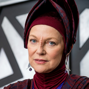 Vedek Winn, personagem de Star Trek: Deep Space Nine, veste a túnica religiosa bajorana em tons de bordô com lenço na cabeça, brincos de pérola roxa e expressão séria, em frente a um fundo cinza desfocado.