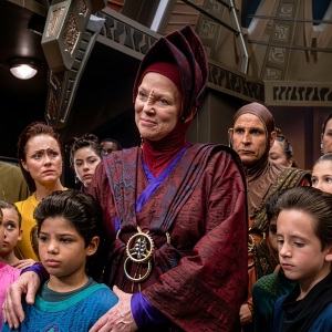 Cena de Star Trek: Deep Space Nine. A candidata a Kai Vedek Winn está ao centro, usando vestes tradicionais bajoranas em tons de vinho com detalhes em azul e um lenço característico da cultura bajorana, com uma medalha cerimonial pendurada ao pescoço. Ela está cercada por várias outras personagens ao redor, em pé, dentro de um cenário de estação ou nave. A expressão de Winn é solene e sóbria.