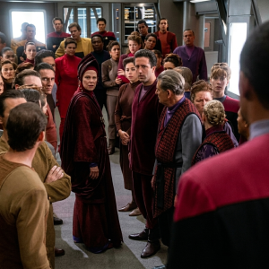 Cena de Star Trek: Deep Space Nine mostrando Kai Vedek Winn, candidata ao cargo de Kai, posicionada no centro, cercada por diversos membros da congregação Bajoriana em vestes religiosas vermelhas e marrom dentro da estação espacial. A personagem está em destaque no meio da multidão, em uma cena de tensão política religiosa.