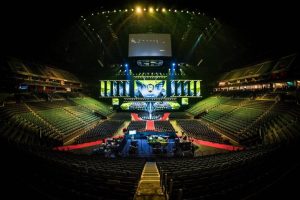 Vista interna da Lanxess Arena em Colônia, Alemanha, mostrando o espaço vazio com assentos organizados em fileiras, um palco iluminado ao fundo e telas grandes suspensas, preparado para um evento. A iluminação destaca a estrutura moderna da arena e o tapete vermelho que se estende pelo chão.